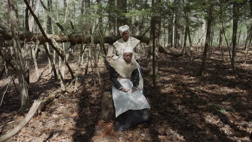 A slave woman is showing another how to slit a throat outside in daylight