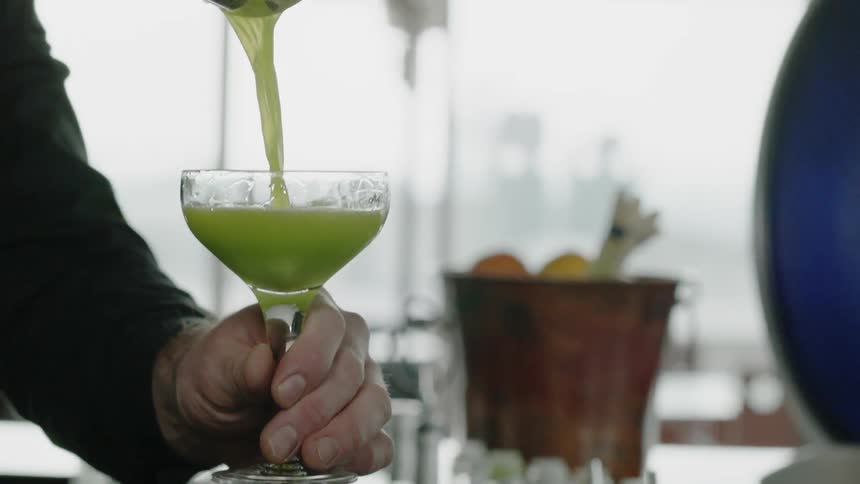 A bartender is pouring a cocktail into a glass at a bar in daylight