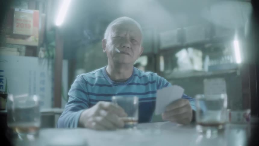 An old man is sitting at a table smoking, drinking, and holding cards