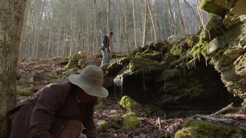 A man searches around in rocks and leaves as a soldier stands watch