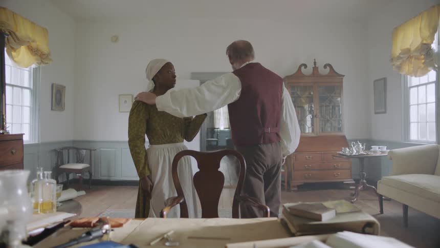 Woman holds a plate out for a man who puts his tea cup on it and pats her shoulder and walks away