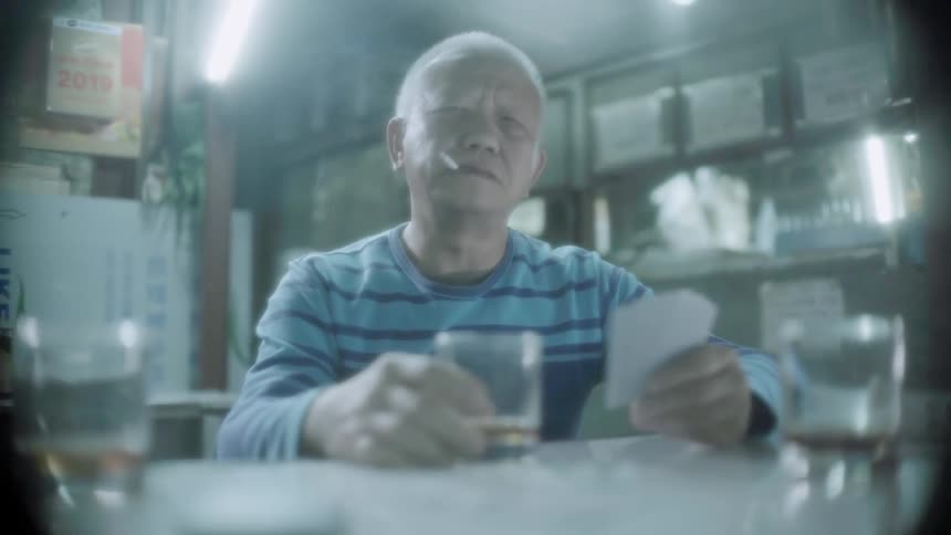 An old man is sitting at a table smoking, drinking, and holding cards