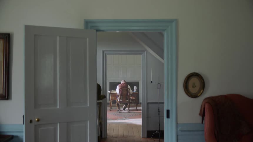A slave woman is carrying a tray through a house to a man at a desk in daylight