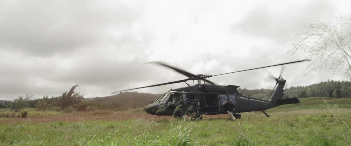 A black helicopter sits in a field with propellers spinning while a man gets out and kneels while carrying a case
