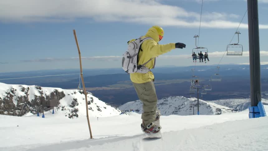 A snowboarding going downhill and jumping as others are shown riding the lift to the top