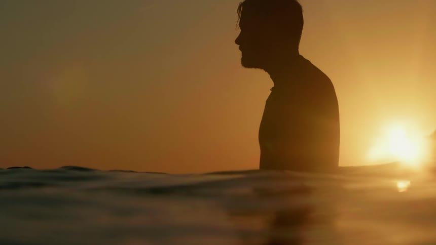 The man is standing waist high in the water at sunset