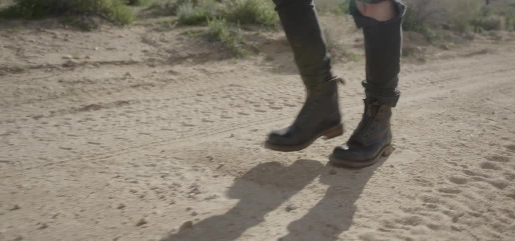 A person wearing boots and torn jeans walks on a dirt road