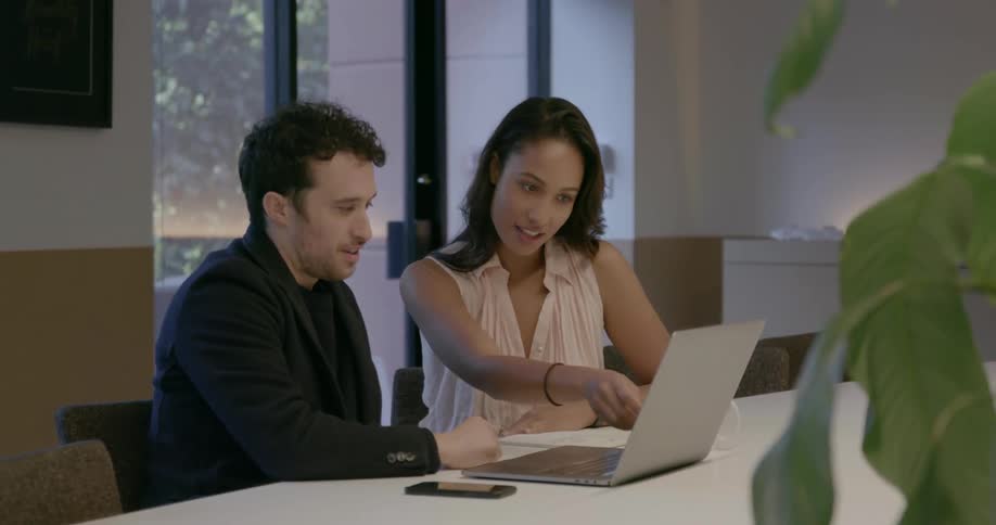 A man and woman sit at a table working on the same laptop