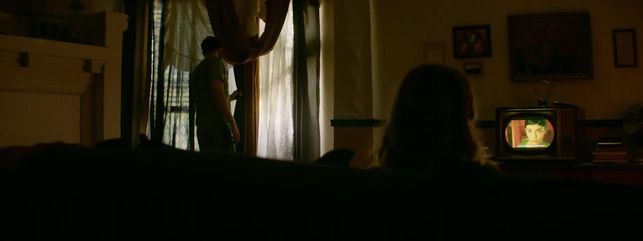 A man peeks out the window as a woman watches television