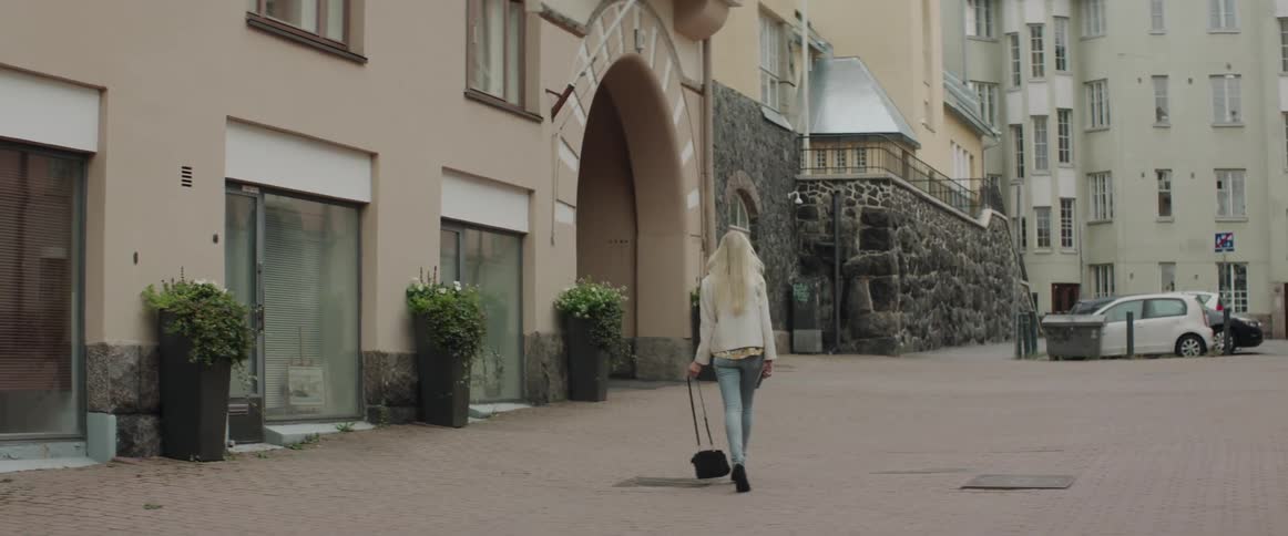 A blonde woman in a white jacket walks along a quiet city street.