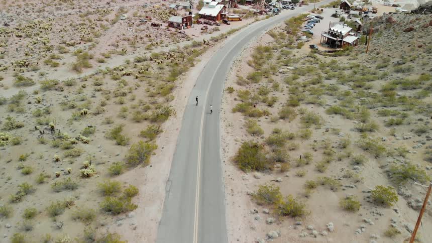Two people skateboard on a rural road leading to town