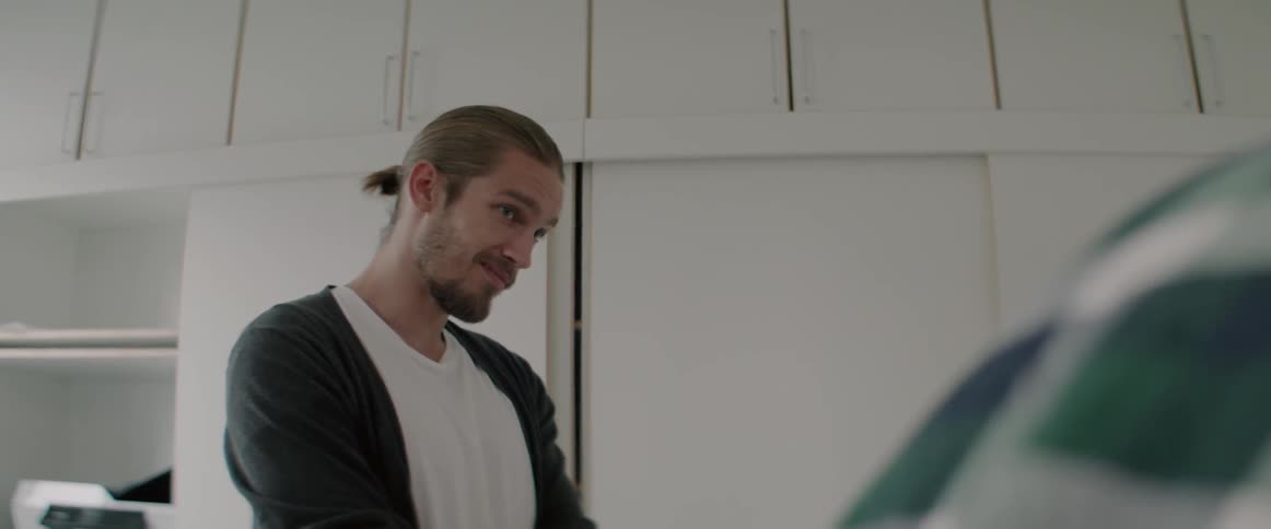 A smiling young man with tied hair looks down while talking to colleagues.