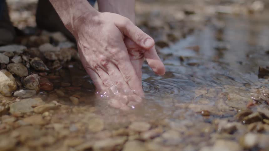 A man is dipping his hands into a stream to wash them on a sunny day