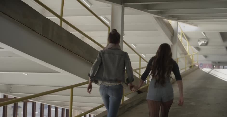 Two women are running hand-in-hand inside a parking garage on a sunny day