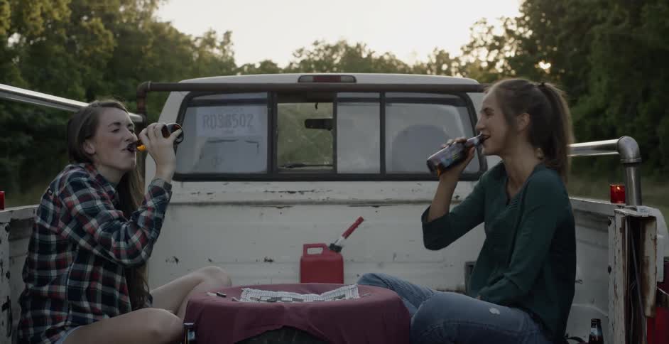 Two women sit in the back of a truck smiling and clinking bottles together then taking a drink
