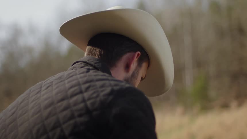 A cowboy is sitting outside in a natural setting deep in thought