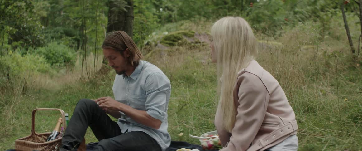 A young man talks to a blonde woman while sitting on a picnic blanket.