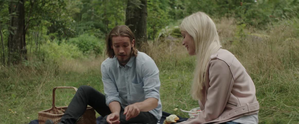 A young man gestures while talking to his blonde girlfriend during an outdoor picnic.