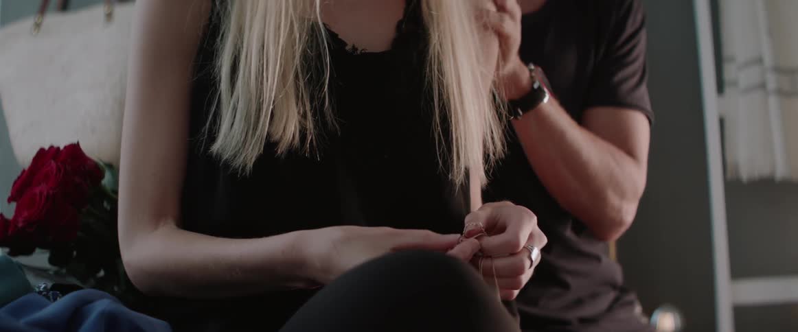 A woman takes off a necklace and hands it to a man behind her
