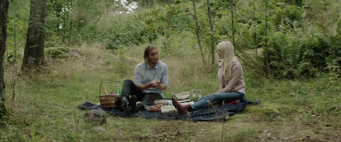 A man and blonde woman sit on a blanket during a forest picnic.