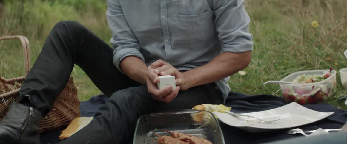 A young man holding a small closed white jewelry box during an outdoor picnic.