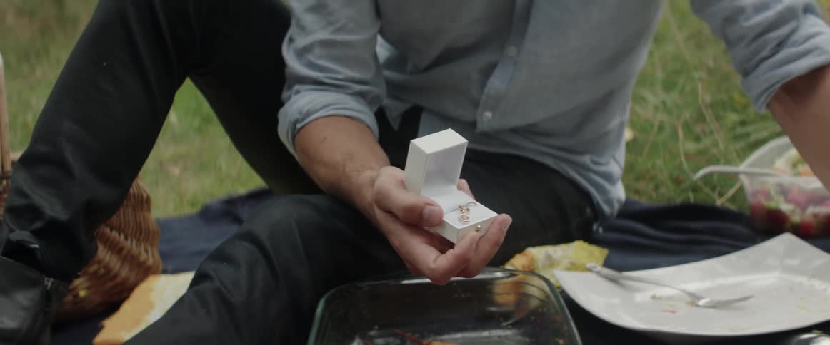 A man is having a picnic with his girlfriend and he opens up a box of jewelry for her