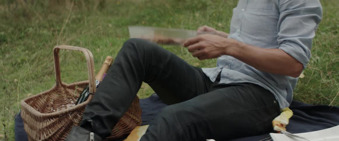 A man reaches for a plastic food container while sitting on a picnic blanket.
