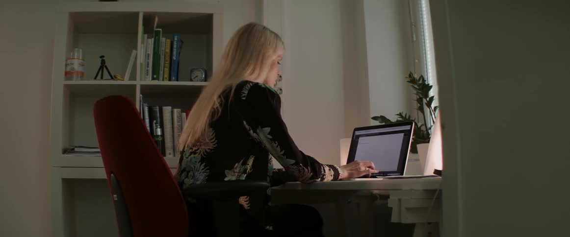 A blonde woman sits in an office chair typing on a laptop at night.