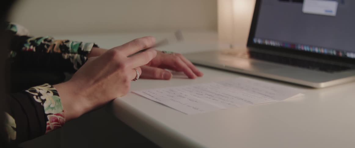 A woman holds a pen at her white desk next to a laptop.