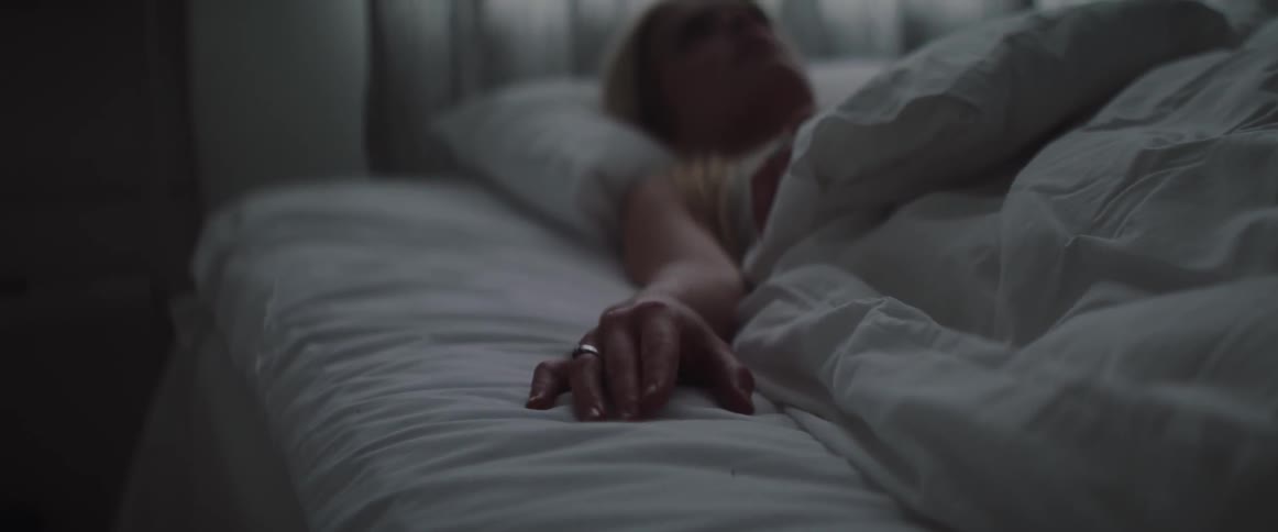 A restless blonde woman lies in bed tapping her fingers on mattress.