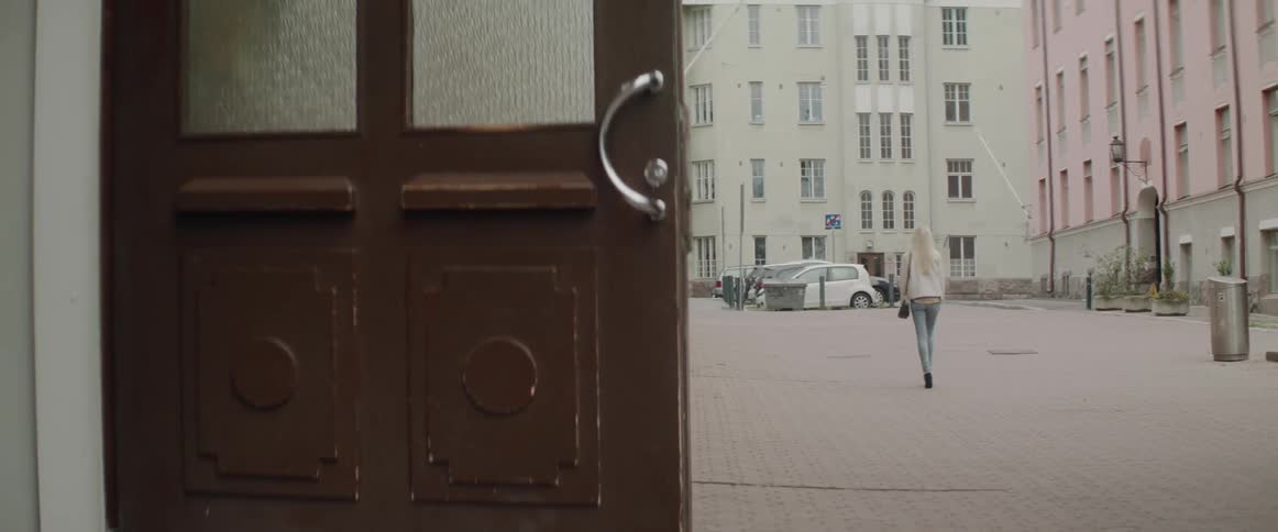 A blonde woman walks down a European city street towards an apartment door.