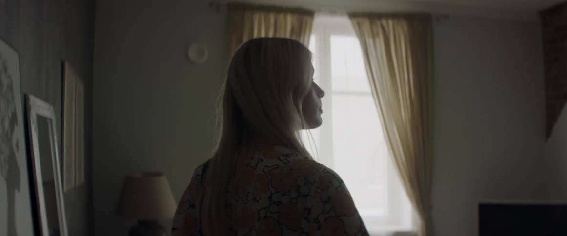 A blonde woman looks around a brightly lit living room near a window.