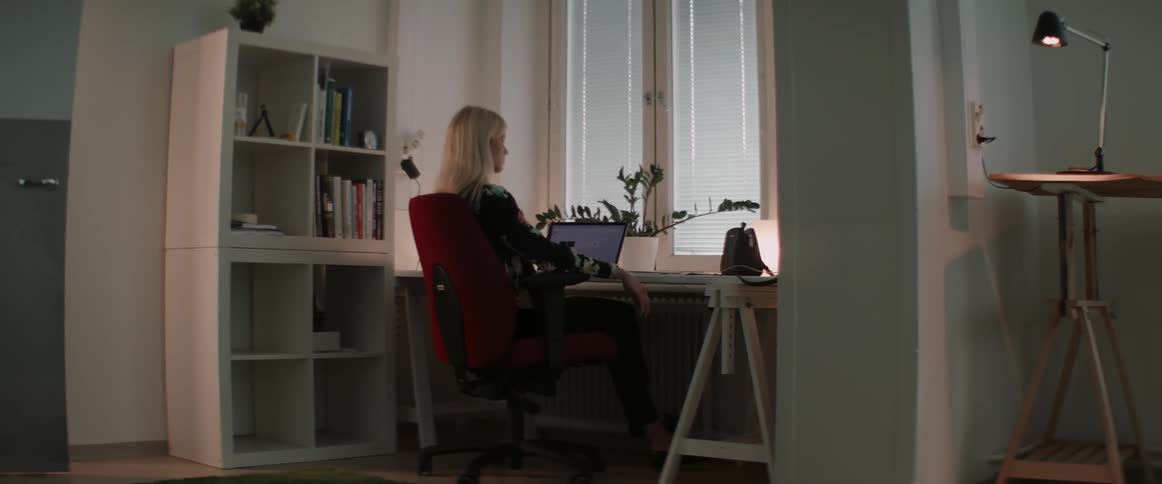 A blonde woman sits at a home office desk working late on laptop