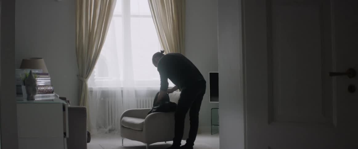 A young man stands by a window in a room packing his black backpack.