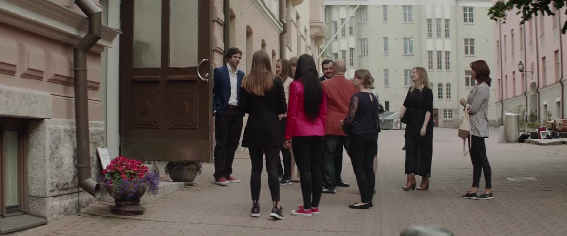 A large group of smartly dressed friends stands and talks outside building entrance.