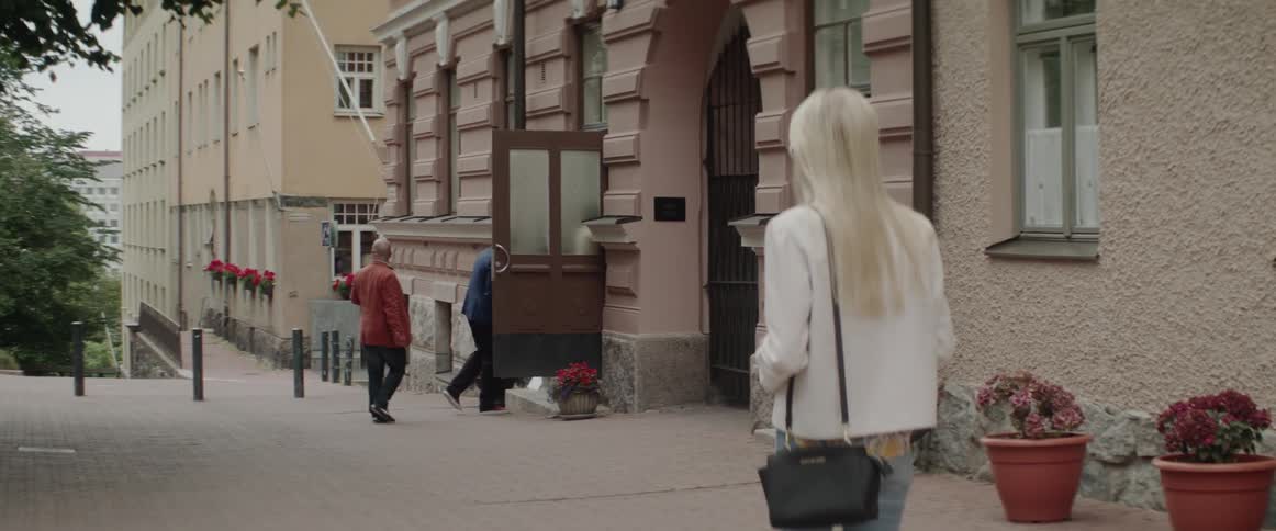 A blonde woman stands on the sidewalk watching people walk into a building.
