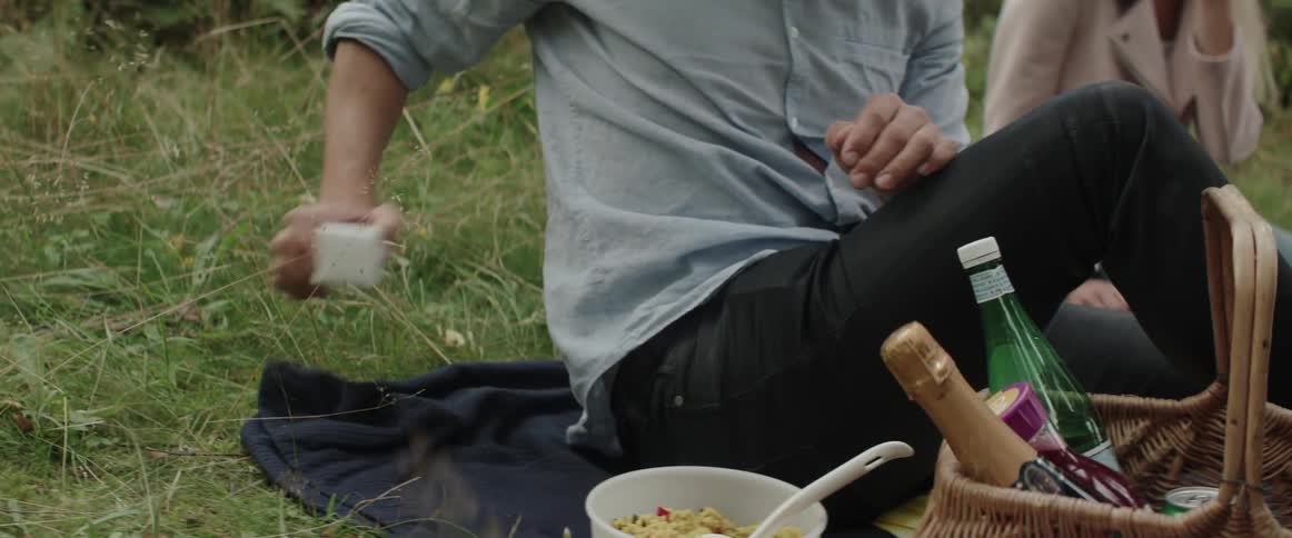 A young man reaches for a small white jewelry box during an outdoor picnic.