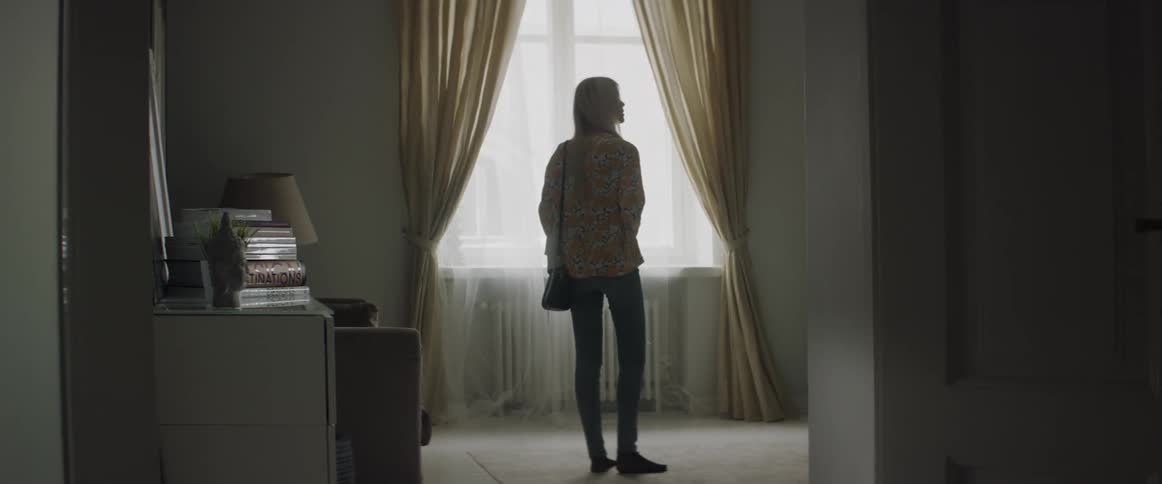 A blonde woman stands in a living room near a window looking around.