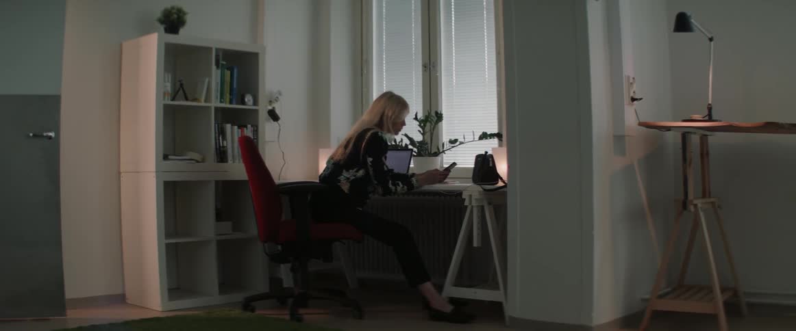 A woman sitting in a red desk chair checks her smartphone while working late.