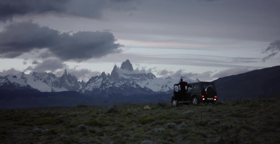 A man stands on the running boards of his suv as he views a mountain landscape at dusk