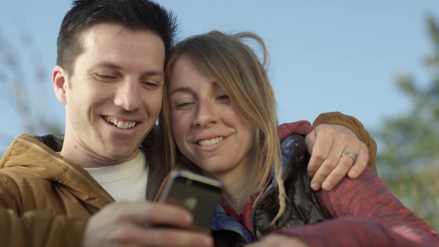 A woman gives a man a small kiss on the cheek while they look at a cell phone