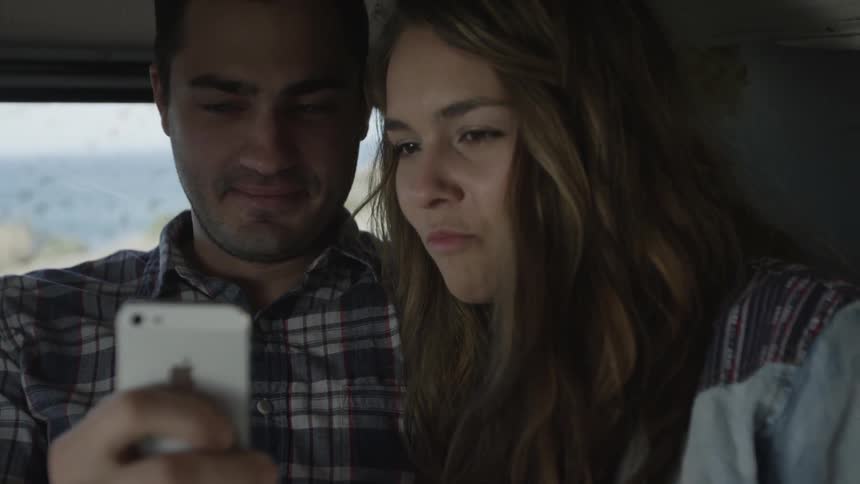 A man and woman sitting in a vehicle, looking at a cell phone and laughing