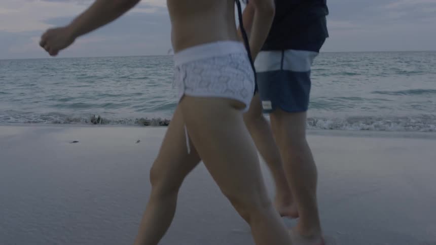 A man and woman walk barefoot on a beach at sunset