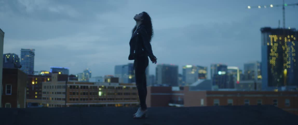 A woman is taking in a deep breath and throwing her arms back outside at dusk