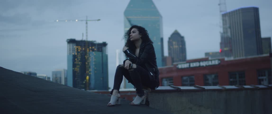 A woman sitting on a rooftop in a city at dusk is putting away her cell phone
