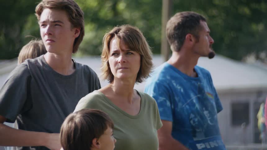 A woman rubs her son's back as they stand in a group in a lot