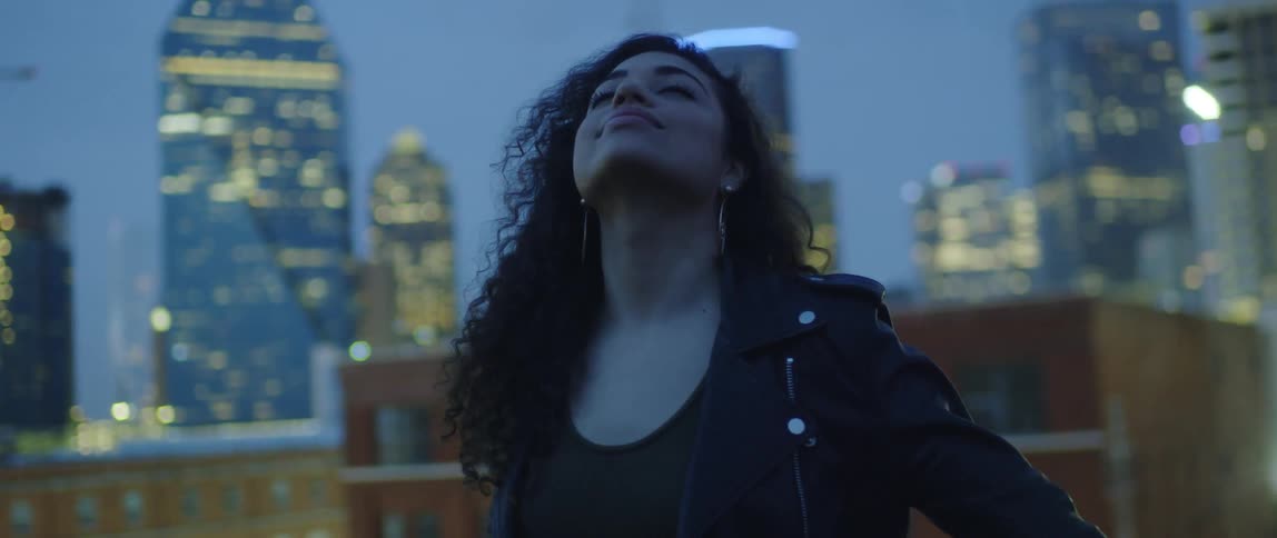 A woman smiles and sways as she looks up at the city sky