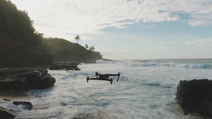 A drone hovers near the water's edge then flies toward the waves