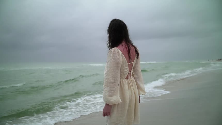 A woman in a white dress walks to the rough waters