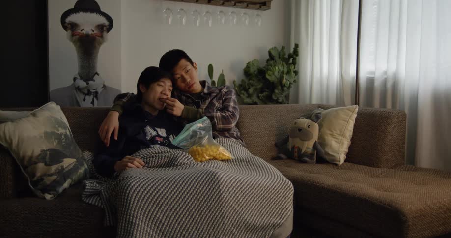 Two men cuddle on a couch and eat snacks